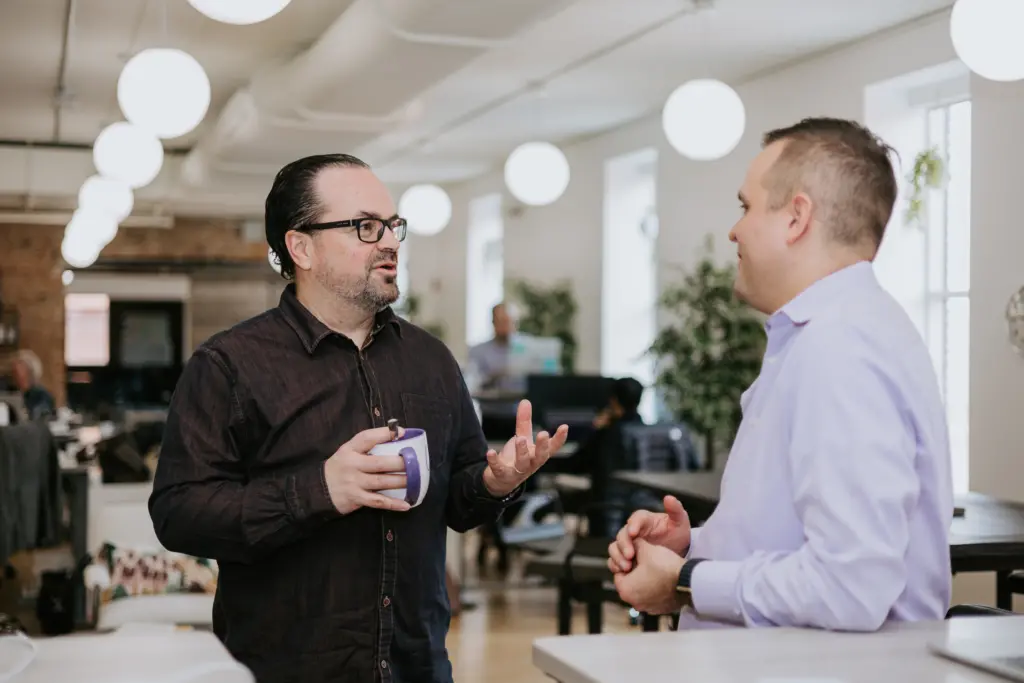 10+ Clear Benefits of Mentoring Programs 4 Two men in an office discussing the benefits of mentoring while at a standing desk.
