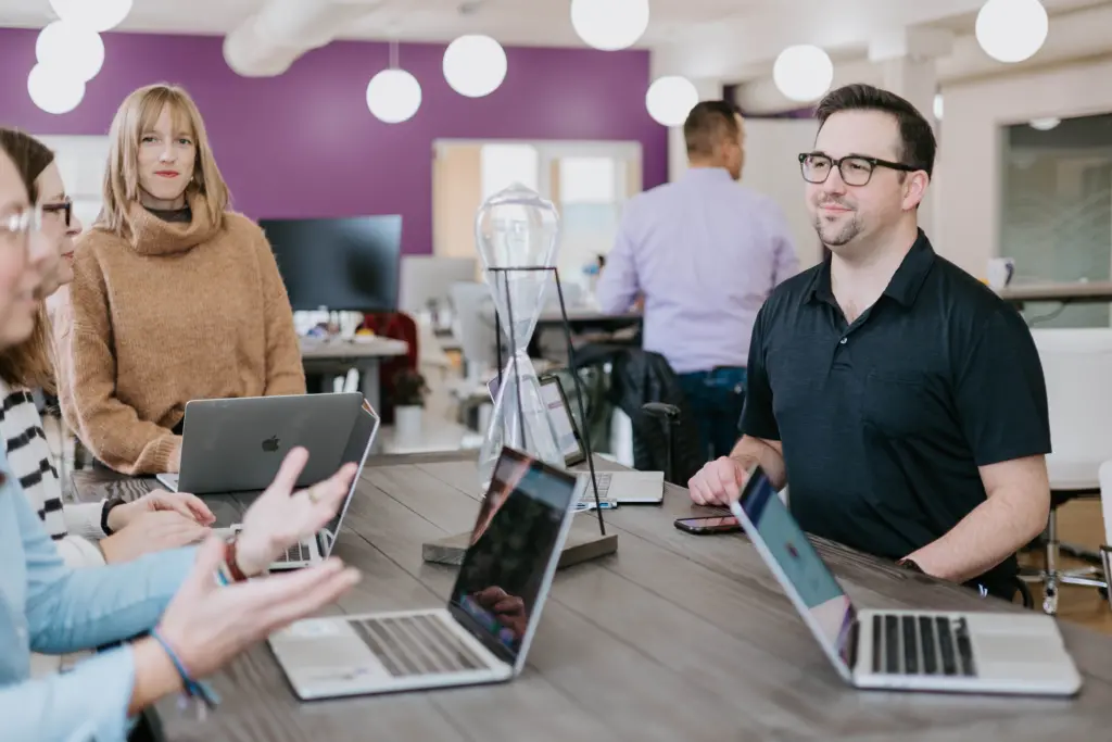 What Is Employee Development? In-Depth Strategy Guide 5 Employees sitting at a long table in an office with purple theme walls engaged in collaborative engagement discussions.