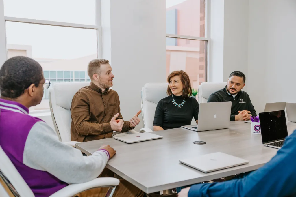 5 Types of Mentoring Program Formats (with Detailed Use Cases) 8 Employees in a group mentoring setting sit around a table in a discussion.