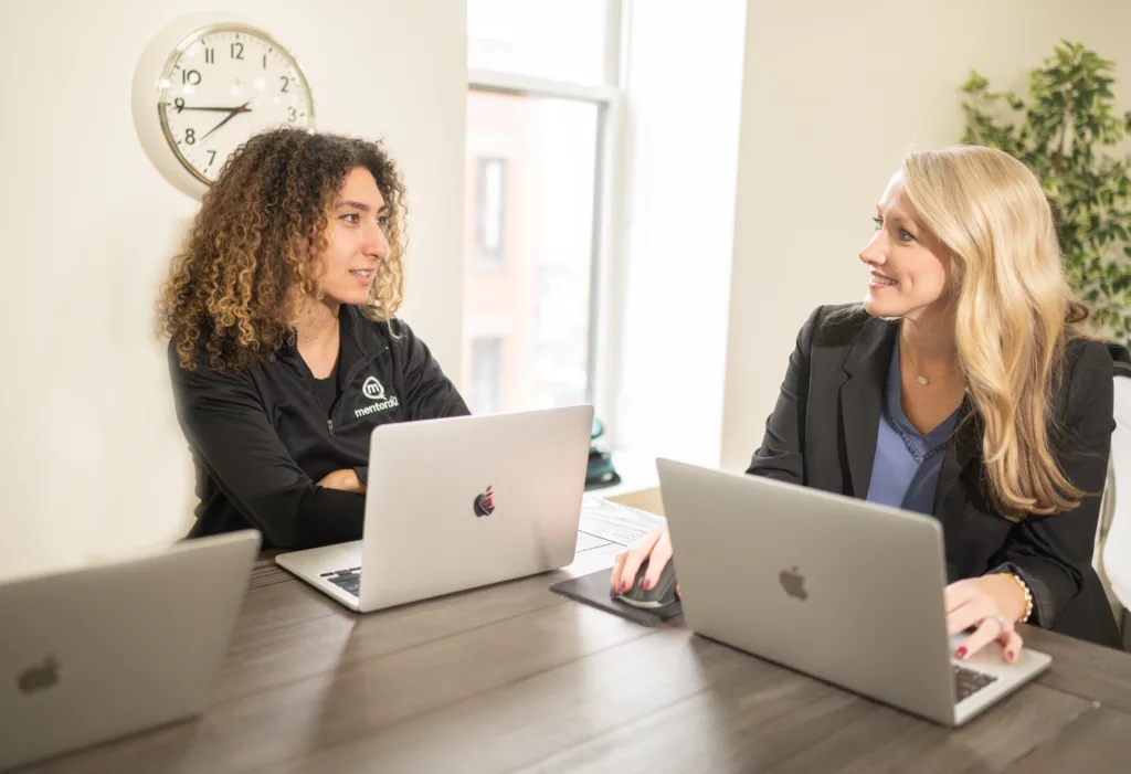 5 Types of Mentoring Program Formats (with Detailed Use Cases) 6 Image of two female employees around a table planning development for different types of mentoring programs.